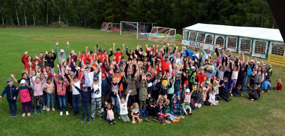 The photo shows the children of the city recreation with supervisors and visitors. They are all standing on the sports field and waving at the camera.