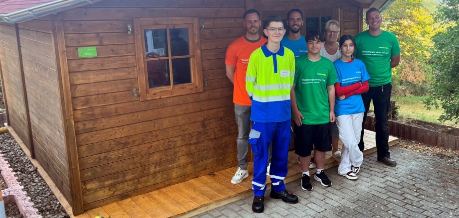 The photo shows seven people standing in two rows in front of the renovated garden shed.
