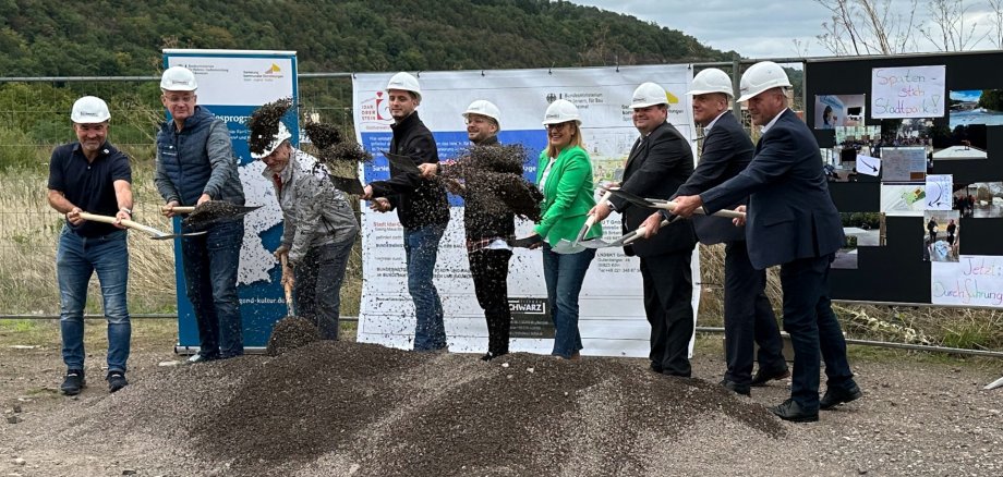 The photo shows the above people from left to right. They are standing at a pile of sand with construction helmets and spades and throwing earth into the air.