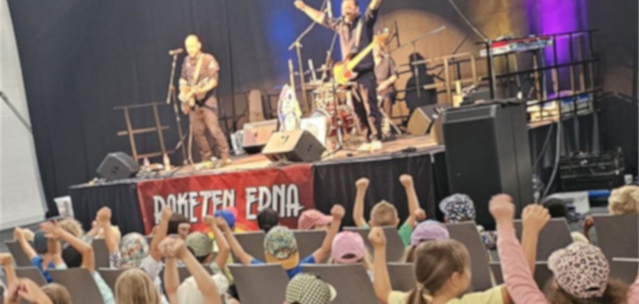 The photo shows lots of children sitting on chairs in front of the stage. The band Raketen is playing on stage,