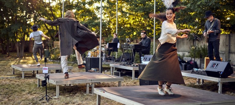 The photo shows three dancers from the company dancing individually on stage platforms outdoors. Behind them are musicians who are also placed individually on stage platforms.