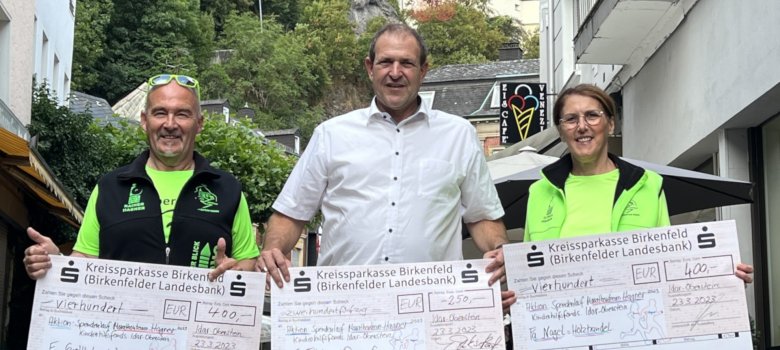 The photo shows, from left, Rainer Hagner, Lord Mayor Frank Frühauf and Ilonka Hagner. They are standing next to each other, each holding a large donation check. The Felsenkirche church can be seen in the background.