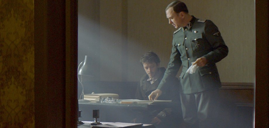 The photo shows two men in a dark room. One is sitting at a desk. The other is standing next to him, leafing through a book lying on the desk.