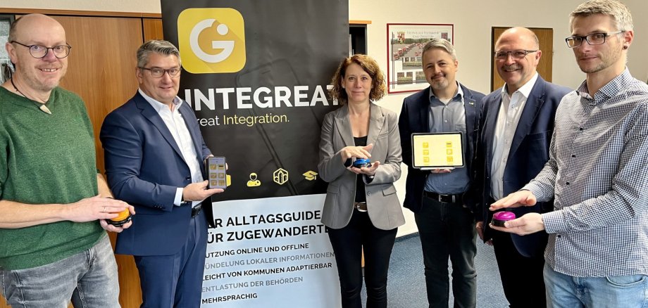 The photo shows six people standing in front of a roll-up with the words Integreat app on it. They are holding buzzers in their hands with which they symbolically activate the app.