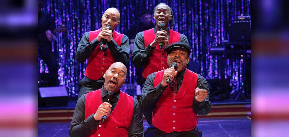 The photo shows four singers sitting two by two on bar stools on the stage and singing.