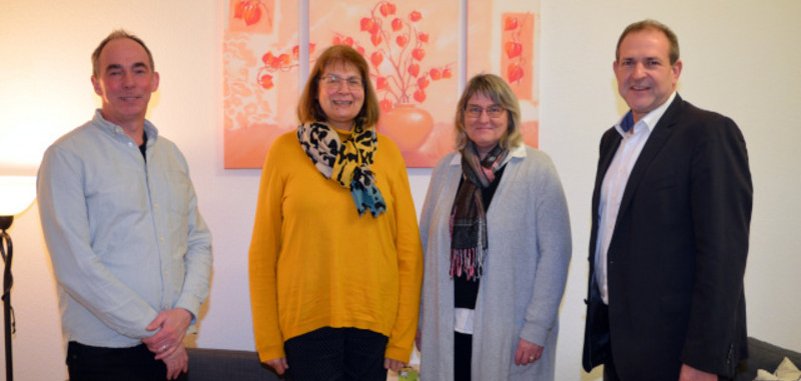The photo shows the aforementioned people standing next to each other in front of a wall decorated with paintings and looking into the camera.