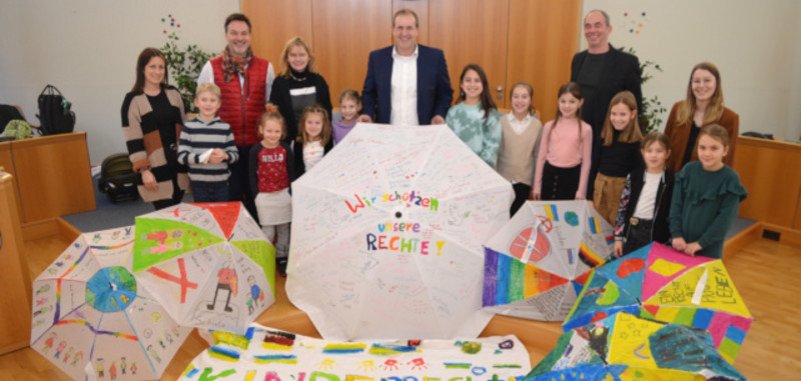 The photo shows Mayor Frühauf, Frank Schnadhorst, Michael Schweizer, the teachers and pupils. They are standing in the conference room behind the stretched umbrellas and the large banner, looking into the camera.