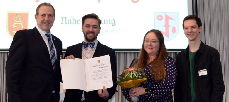 The photo shows the people mentioned. They stand next to each other on the stage and look into the camera. Moritz Forster presents the certificate of appointment.