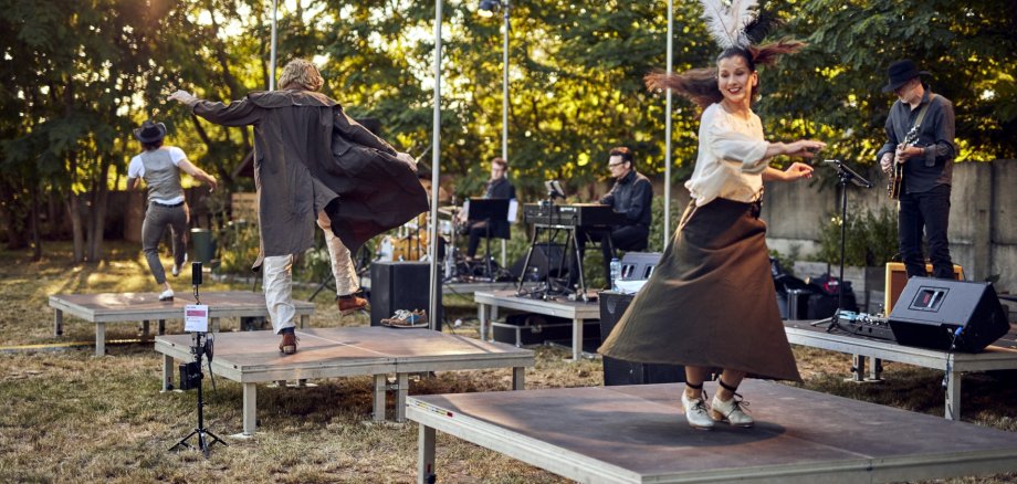 The photo shows three dancers from the company dancing individually on stage platforms outdoors. Behind them are musicians who are also placed individually on stage platforms.