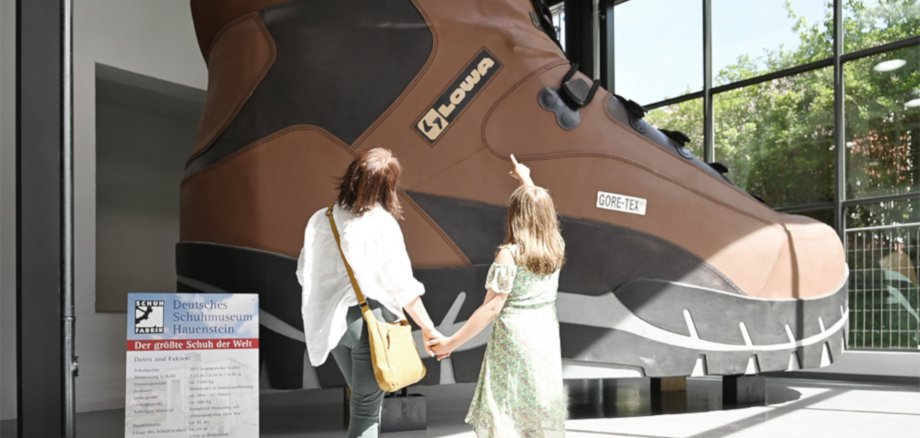 The photo shows two visitors standing in front of the largest shoe in the world at the Hauenstein Shoe Museum.