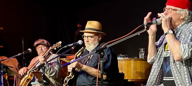 The photo shows the three musicians sitting next to each other on a stage and playing their instruments.