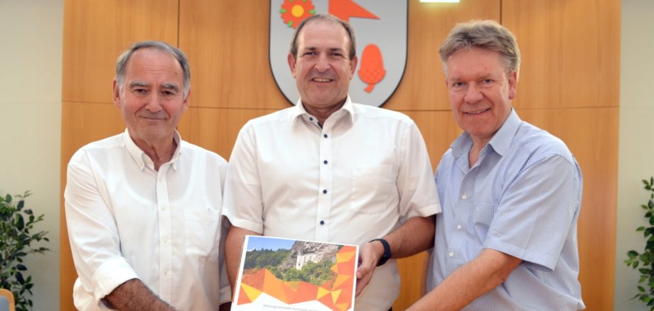 The photo shows, from left to right, District Councillor Peter Simon, Alexander Hauck and Lord Mayor Frank Frühauf holding one of the education folders up to the camera.