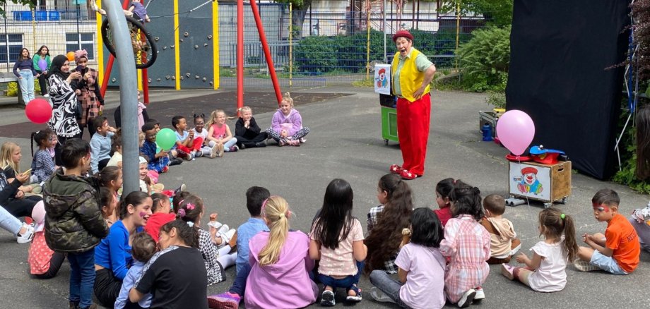 The photo shows a large group of children sitting in a semi-circle in front of the clown Kuni on the event site.