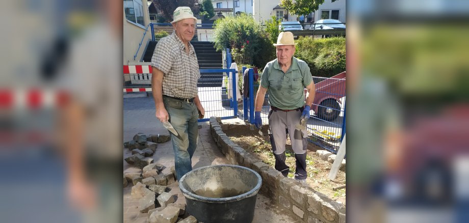The photo shows two members of the Open Group in work clothes. They are standing by the wall they are building.