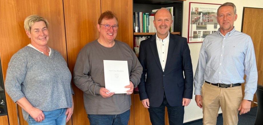The photo shows, from left, Marion Müller, Rudi Bonn, Mayor Friedrich Marx and Christoph Hahn (Staff Council representative). They stand in a row in front of a wall unit and look into the camera. Rudi Bonn holds the certificate of thanks in his hands.
