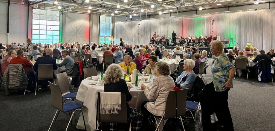 The photo shows a view of the Idar-Oberstein trade fair. The numerous guests are seated at round, laid tables. In the background you can see a stage on which a music band is playing.