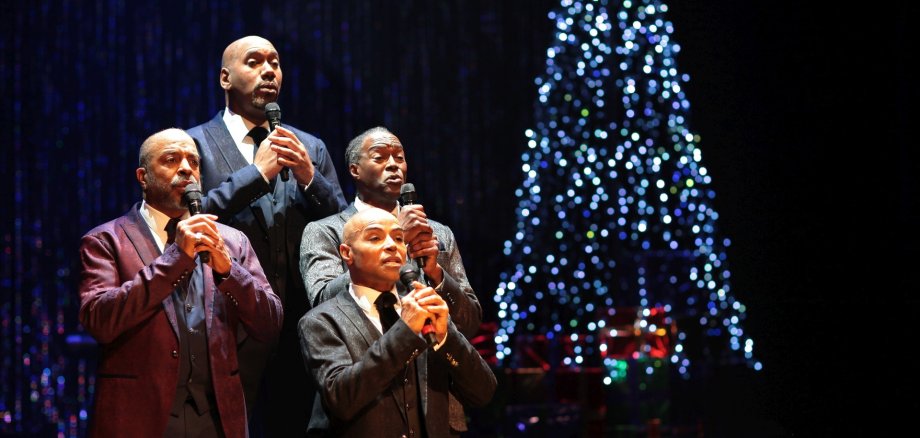 The photo shows four singers standing in front of a Christmas tree.