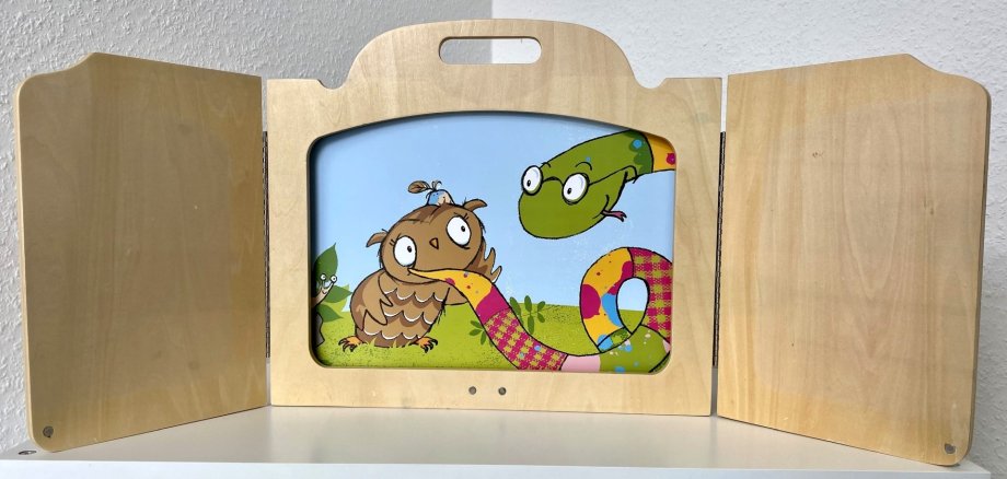 The photo shows a wooden Kamishibai frame. It stands on a table, in the frame is a picture card from the book "The owl with the bump".
