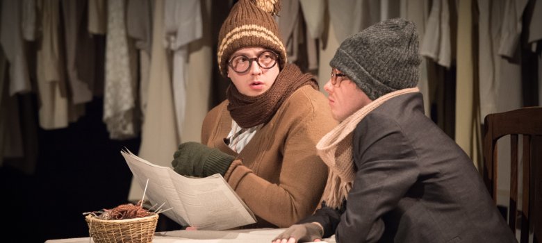 The photo shows two performers sitting at a table wearing winter jackets and hats.