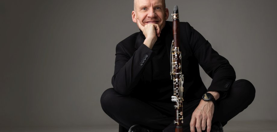 The photo shows the clarinettist sitting cross-legged on the floor in a black outfit. His instrument is on the floor in front of him.