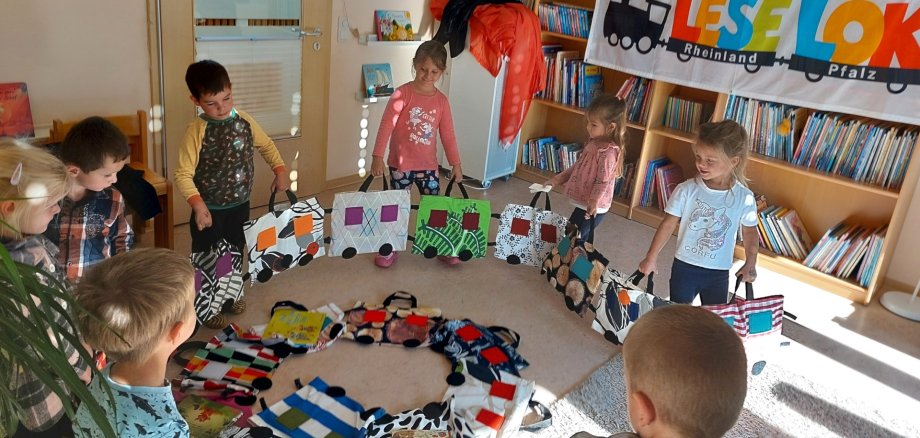 The photo shows a view of the daycare center's reading room. Some children are kneeling in a circle around the train and holding each of the two wagons in their hands. A banner with the inscription "Lese-Lok" hangs on the bookshelf in the background.
