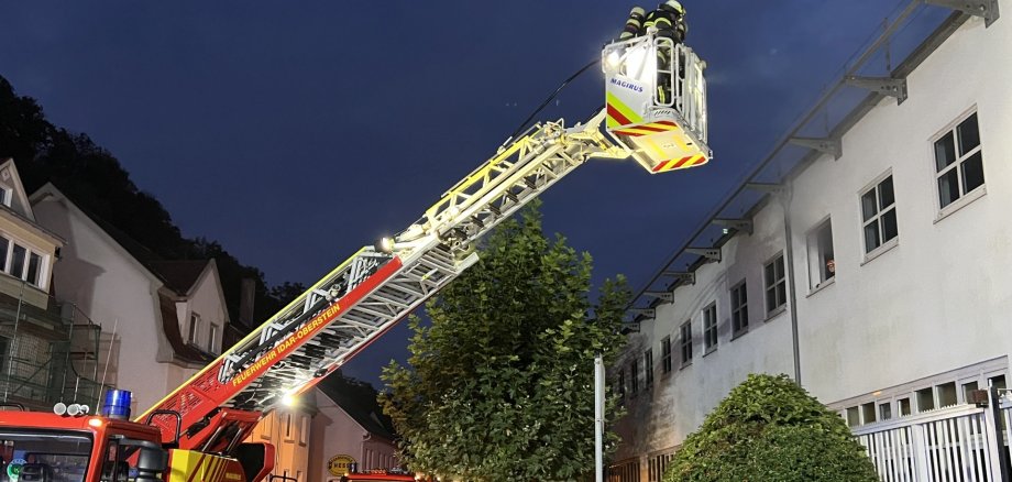 The photo shows two illuminated fire engines in front of the OIE building as darkness falls. One of them is the turntable ladder with two firefighters going up. Another firefighter is walking towards the building.