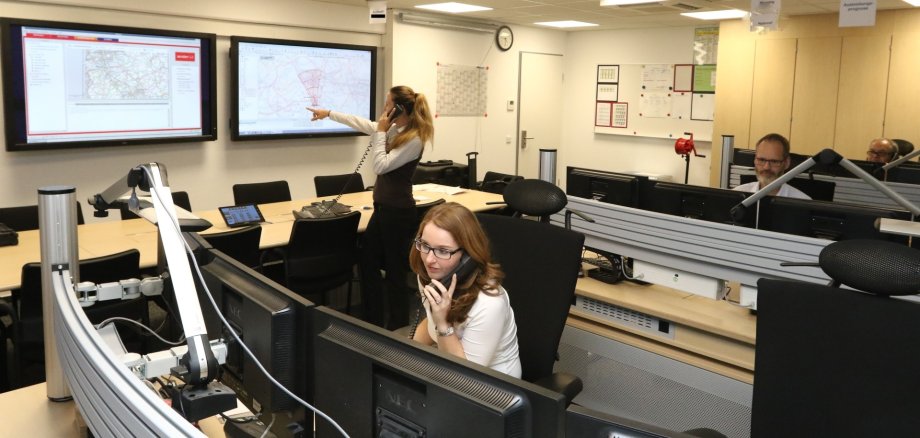 The photo shows a view into the BBK warning center, a woman sits in front of a screen and talks on the phone, a woman talks on the phone and points to a large screen on the wall, a man looks at his screens.