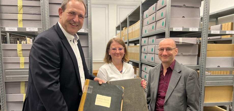 The photo shows the aforementioned people standing in front of the shelves in one of the stacks. Mayor Frühauf and Dr. Müller hold historical books in their hands.