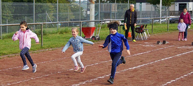The photo shows three children running on the track. The timekeepers and two spectators can be seen in the background.