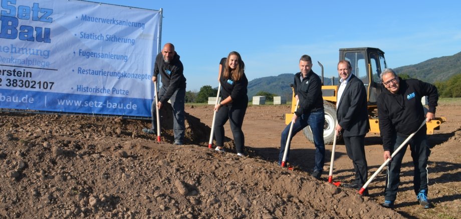 The photo shows the people mentioned. They are standing in a row on a mound of earth with spades in their hands. The spades are digging into the earth. A Setz construction company banner can be seen on the left-hand side of the picture.