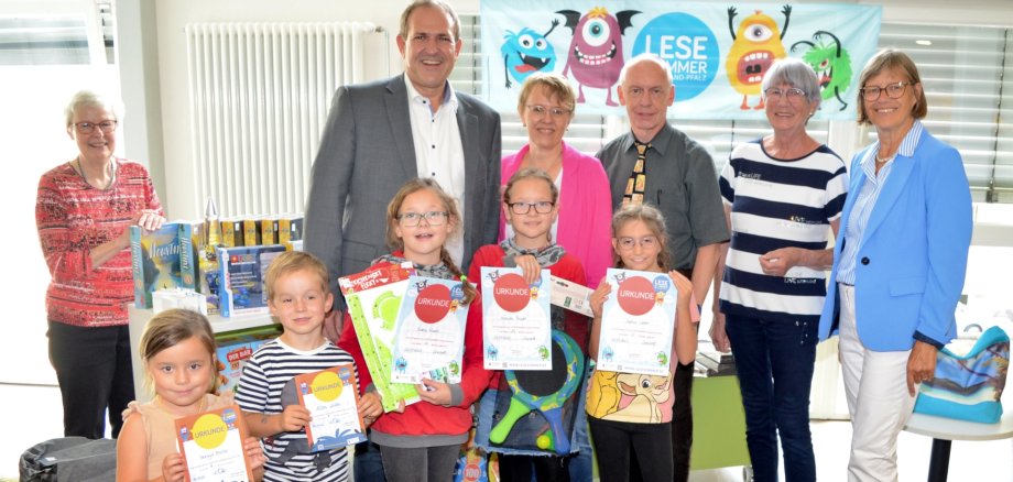 The photo shows the people mentioned. They are standing in front of a table with raffle prizes in the city library together with some children holding their certificates.