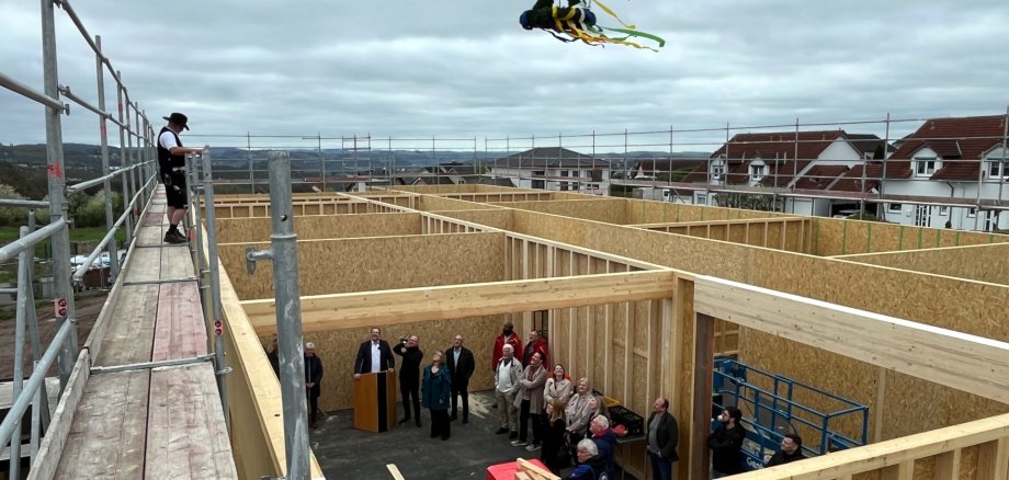 The photo shows a view of the construction site from above. On the left on the scaffolding is the carpenter giving the topping-out speech, below in the building are the guests of the topping-out ceremony looking up at the carpenter.