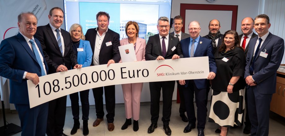 The photo shows the people mentioned. They are standing next to each other in a meeting room at the hospital. They are holding a long banner with the amount 108,500,000 euros and the Idar-Oberstein Clinic logo printed on it.