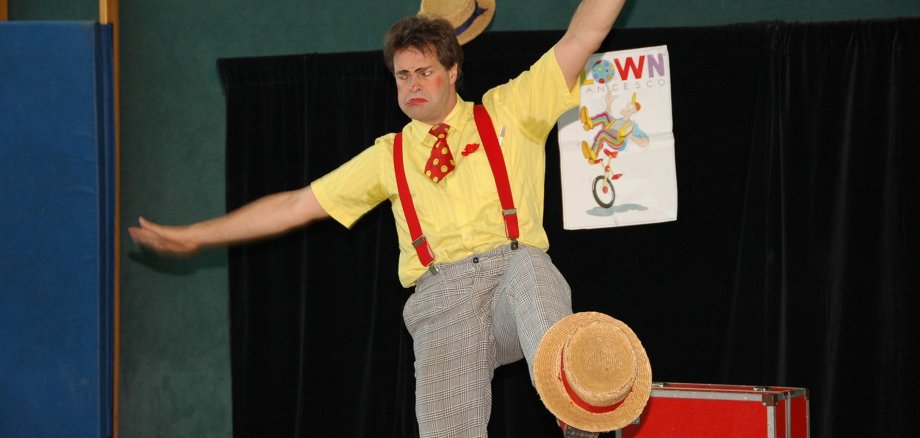 The photo shows a man in clown costume balancing on his right leg and juggling a hat at the foot of his left leg.