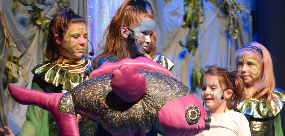 The photo shows four young actresses. They are wearing colorful make-up and imaginative costumes. One of the girls is holding the fish of life (a large plush fish) in her hands.