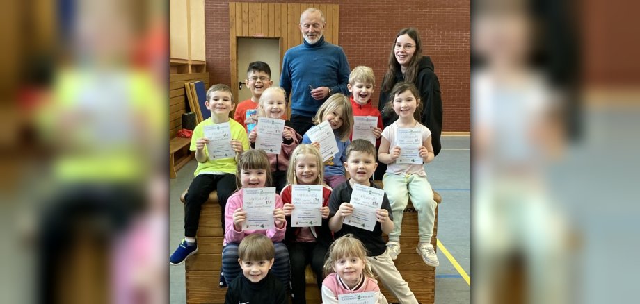 The photo shows eleven children, Klaus Juchem and an educator. The children are sitting in the sports hall in front of and on a box (the sports equipment) and are holding certificates in their hands. The adults are standing behind them.