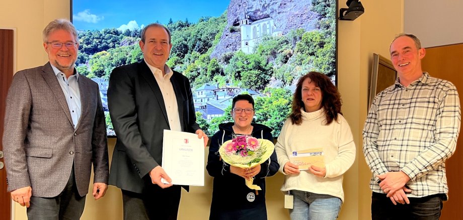 In the picture, senior civil servant Wolfgang Petry, Mayor Frank Frühauf, Petra Forster, Chairwoman of the Staff Council Susanne Becker and Head of the Youth Welfare Office Michael Schweizer (from left) stand next to each other and look into the camera. Mayor Frühauf holds the certificate of thanks, Petra Forster a bouquet of flowers. Behind them hangs a large screen on which a photo of the Felsenkirche can be seen.