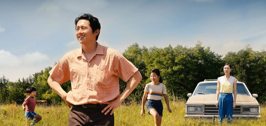 The photo shows a family of four, the parents with a daughter and a son. They are standing in a meadow with tall grass, behind them is their car. Father and mother are looking into the distance, the children are jumping around.