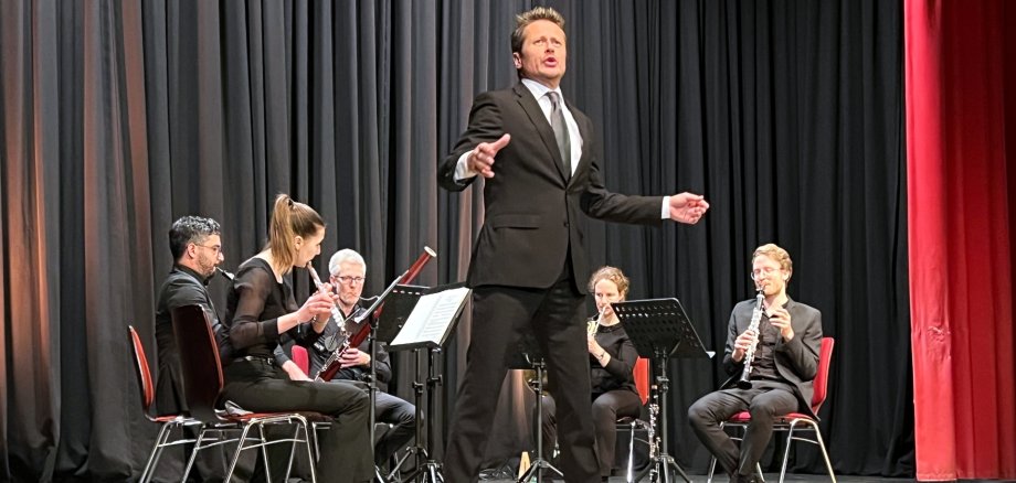 The photo shows the actor Roman Knižka standing at the front of the stage of the Stadttheater and reciting a text. Behind him, the OPUS 45 ensemble sits in a semi-circle on chairs and plays music.