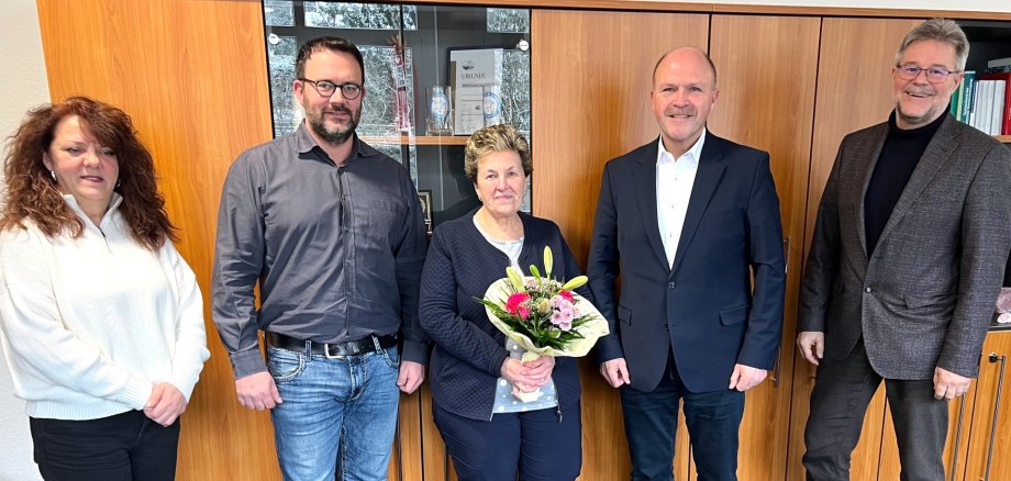 The photo shows, from left to right, the Chairwoman of the Staff Council Susanne Becker, Christoph Alt from Facility Management, Irene Herrherdt, Mayor Friedrich Marx and the Head Clerk Wolfgang Petry. They are standing in front of a wall unit and looking into the camera, Ms. Herrherdt is holding a bouquet of flowers in her hands.