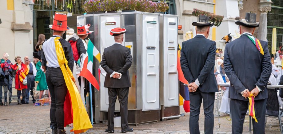 The photo shows disguised people standing outside in a square in front of a toilet facility.