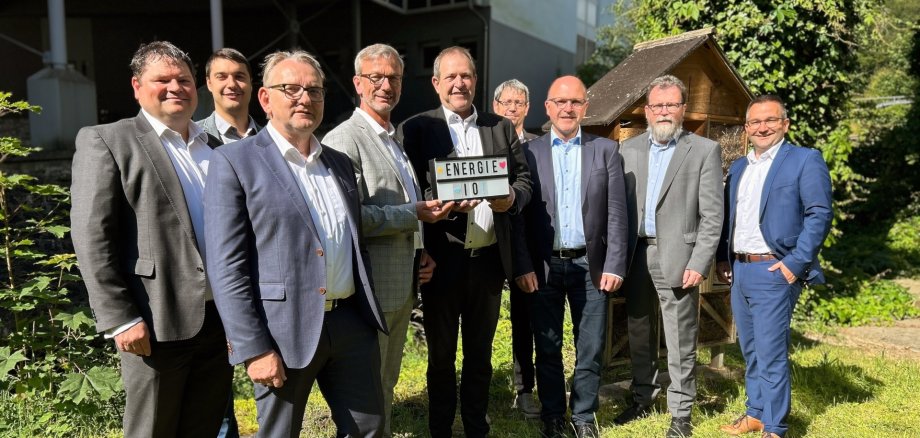 The photo shows the people mentioned in the text. They are standing next to each other on a meadow and looking into the camera. Mr. Johann and Mayor Frühauf are holding a sign with the inscription Energie IO.