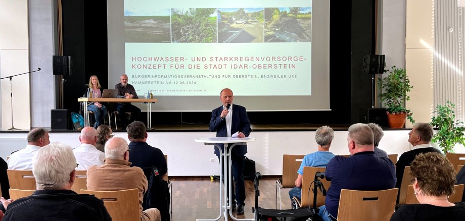 The photo shows a view from the audience area to the stage. Mayor Marx is standing at a high table in front of the stage and is giving a speech. On stage, Mr. Webler and Ms. Baron are sitting at a table next to the screen showing the presentation on the HSVK.