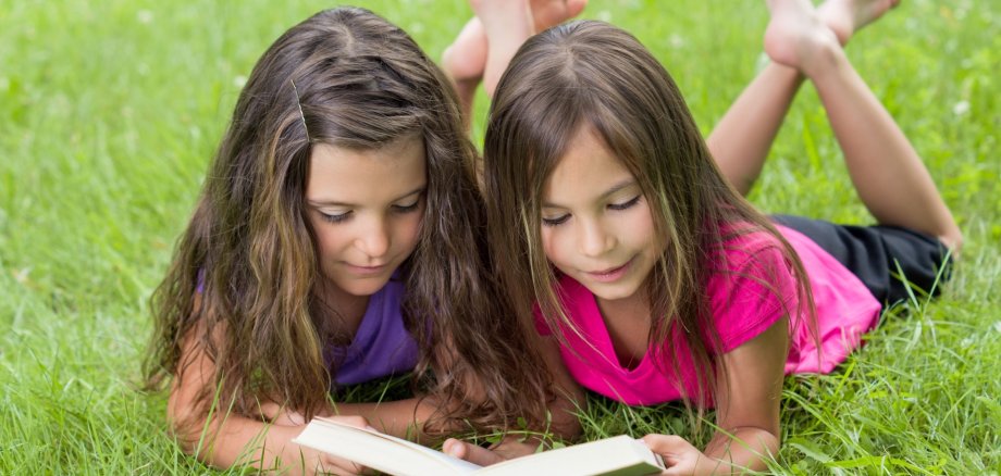 The photo shows two girls lying on a meadow and reading a book together.
