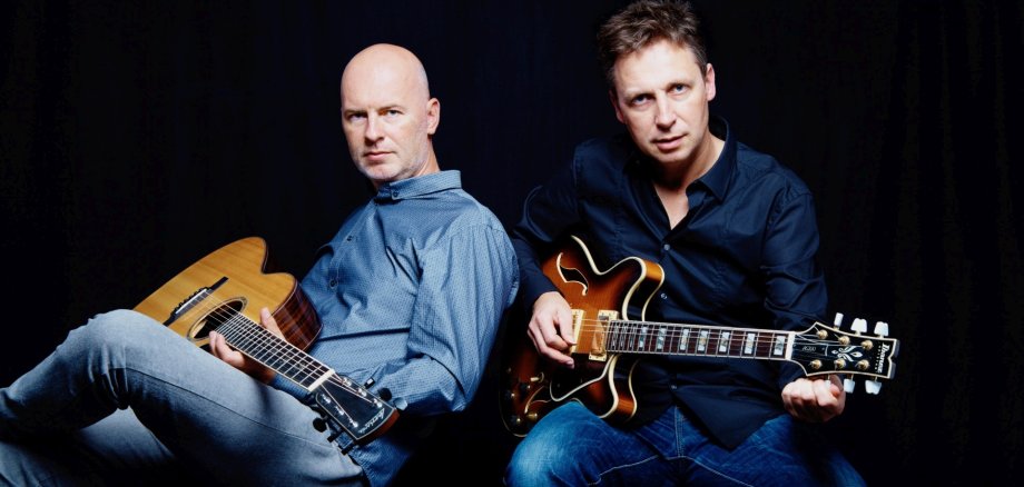 The photo shows two guitarists sitting next to each other on chairs with their instruments and looking into the camera.