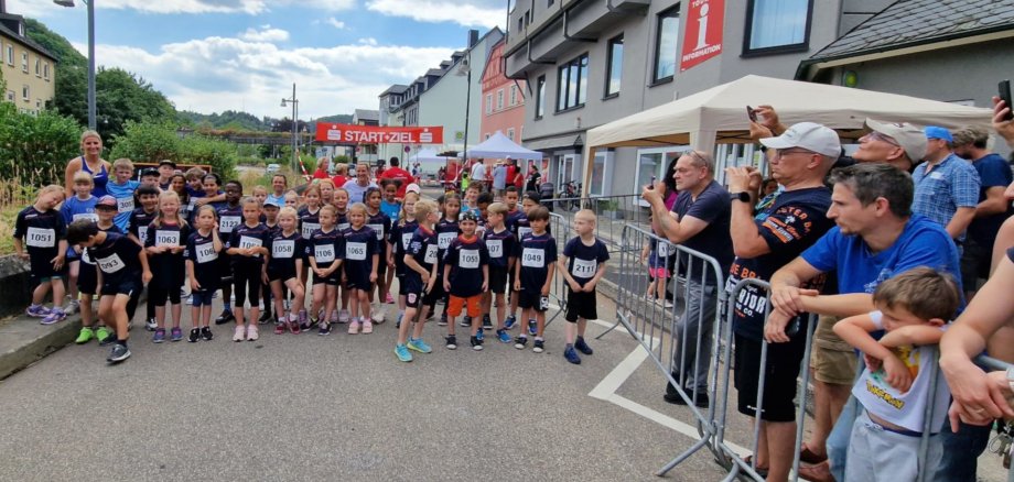 The photo shows many children with start numbers standing in the start-finish area waiting for the starting signal. To the side you can see parents with cell phones taking photos.