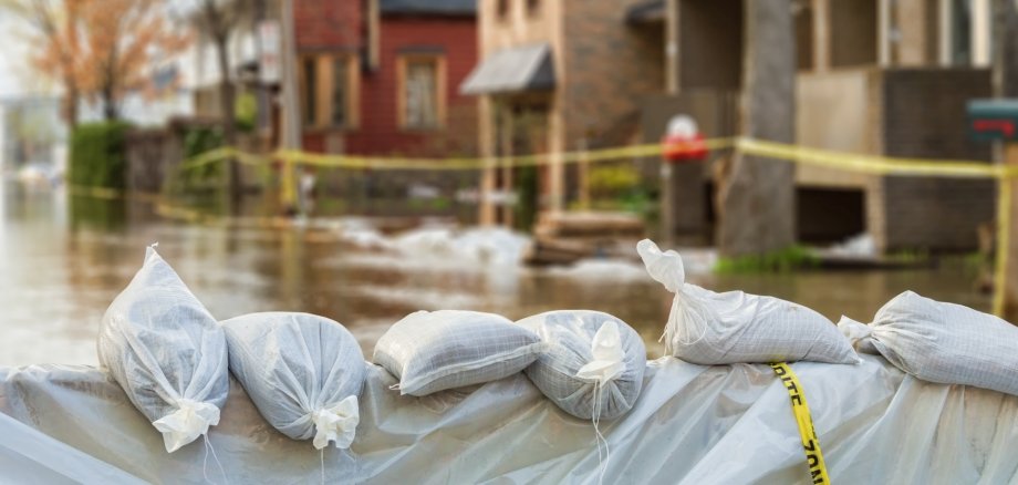 The photo shows a dam made of sandbags in the foreground, in the background you can see blurred houses that are in flood water and are cordoned off with barrier tape.