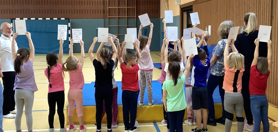 The photo shows many children standing on a large mat in a gym. They turn their backs to the camera and hold up their certificates.