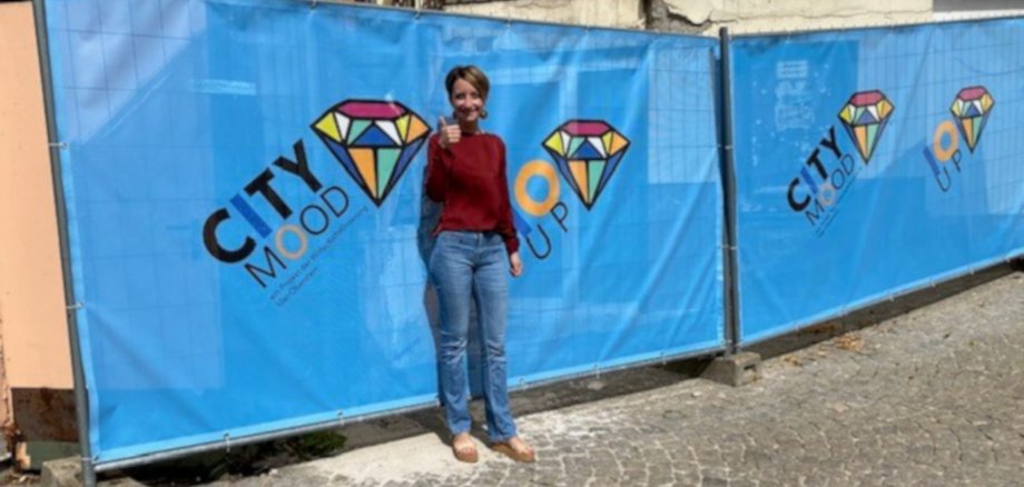 La photo montre la city manager Samira Brächer. Elle se tient debout, le pouce levé, devant deux grandes bannières portant les inscriptions "City Mood" et "IO UP". Les bannières masquent la vitrine du nouveau pop-up store.