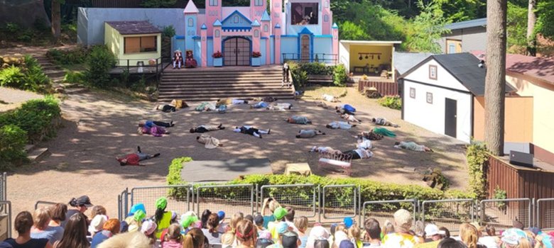 La photo montre une vue de la scène en plein air depuis les sièges des spectateurs. Les acteurs sont tous allongés sur le sol.